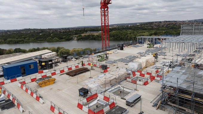 Aerial view of a construction site beside a river, showing a tower crane, steel framework installation, construction materials, temporary barriers and site equipment across a large concrete deck.