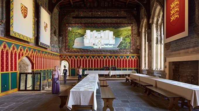 The grand interior of Caerphilly Castle’s Great Hall featuring long banquet tables, colorful medieval-style wall panels, and a large mural of the castle.
