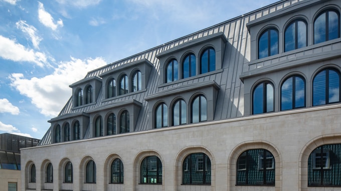 Detail of a contemporary office building showing a grey mansard roof, dormer windows and a light stone façade with arched windows beneath a blue sky.