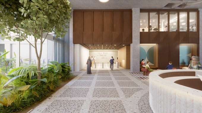 A CGI rendering of a contemporary building entrance and lobby space with extensive planting, including trees and tropical greenery, alongside a terrazzo-style walkway. The interior features high ceilings, timber wall finishes, seating areas and a reception zone. Several people are visible throughout the space, and a sign on the rear wall reads "Fresh Wharf Travel Hub Lifts". Natural daylight enters through large glazed façades, creating a bright and open atmosphere.