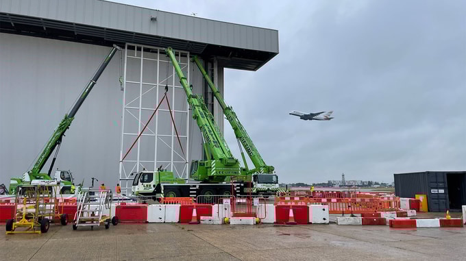 Two green cranes work on a hangar, while a British Airways plane approaches for landing in the cloudy sky above.