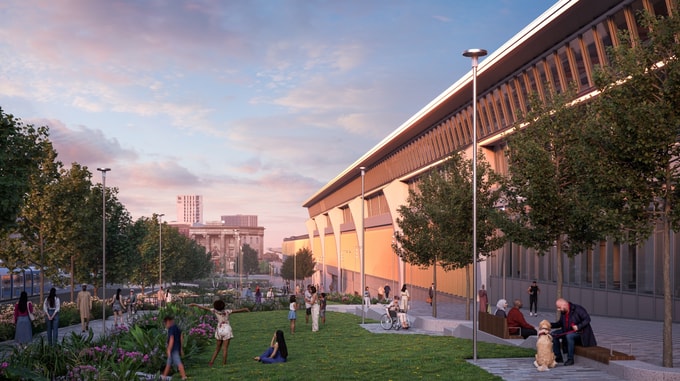 Landscape view of the HS2 Curzon Street station exterior at sunset, showing the long façade with tall windows and structural columns, alongside a landscaped green space where people are sitting, walking, and relaxing among trees and planted areas.