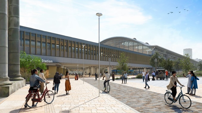 Street-level view of the HS2 Curzon Street station, showing a long façade with tall vertical windows and a curved roof, with people walking and cycling through a paved public space surrounded by trees and landscaping.