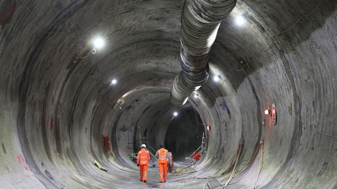 Interior view of a tunnel under construction at HS2 Euston, showing a curved concrete lining, overhead ventilation ducting, lighting, cabling, and two workers in high‑visibility clothing.