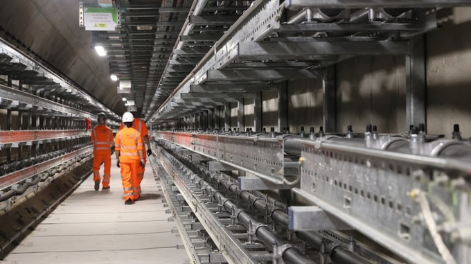 Interior view of an HS2 Euston tunnel showing mounted services, cable trays, and two workers in high‑visibility clothing walking along the tunnel corridor.
