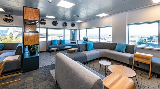 A contemporary office lounge with grey sofas, blue cushions, and wooden tables, set against large windows that reveal a cityscape view.