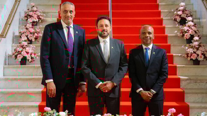 Three men in suits stand on a red carpeted staircase adorned with pink and white flower arrangements on both sides