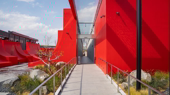 A concrete pathway with metal railings leads between bold red geometric buildings and landscaped gardens under a blue sky