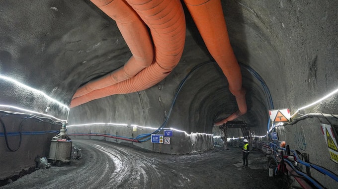 An aerial view of a construction site carved into a green hillside beside a body of water, showing access roads, temporary buildings, excavation areas, and tunnel entrances within the surrounding landscape.