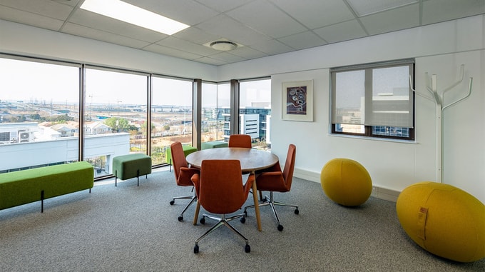 A bright corner meeting space with a round table, orange chairs, green bench seating, large windows overlooking city buildings, and two yellow exercise balls.