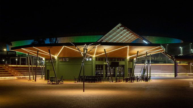 A night view of a pavilion-style building with a slanted roof and warm linear lighting, surrounded by an open brick-paved courtyard.