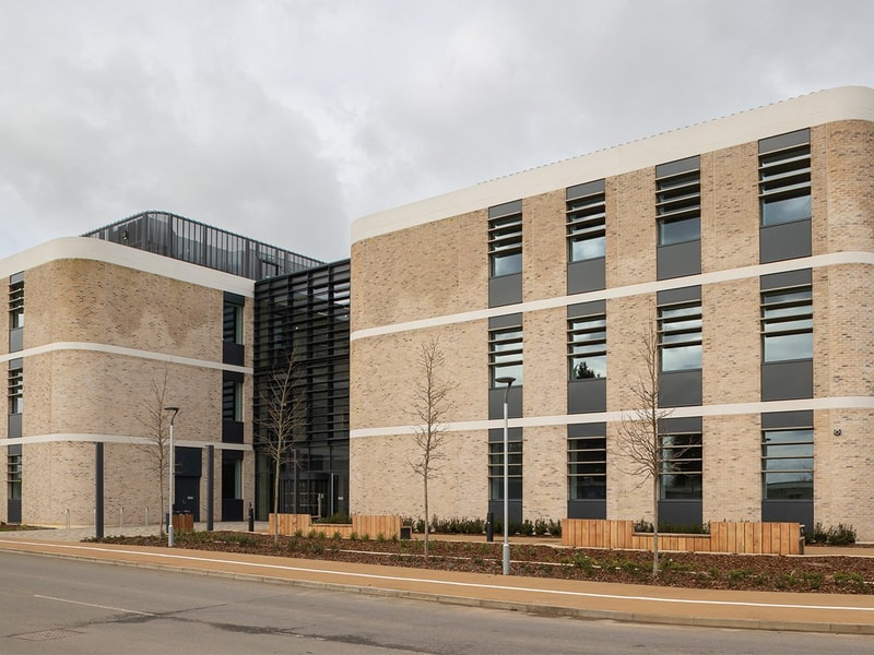 Modern commercial or institutional building with light brickwork, large vertical windows, and horizontal black louvers. The structure features rounded corners, white horizontal bands between floors, and a landscaped frontage with young trees and wooden fencing under an overcast sky.