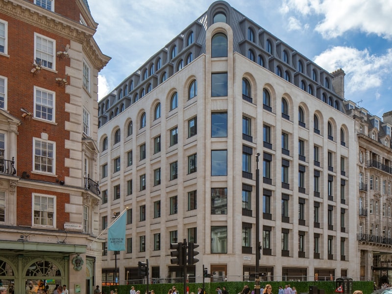Modern office building with a light stone façade and arched windows on a prominent street corner, surrounded by historic buildings in central London. Pedestrians, traffic lights and passing vehicles line the busy street beneath a blue sky with scattered clouds.