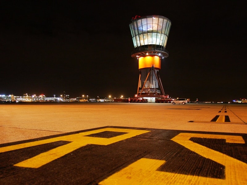Night-time view of an airport runway with large yellow runway markings in the foreground. In the distance, an illuminated air traffic control tower sits on a large transport platform, with airport buildings and lights stretching across the horizon beneath a dark sky. The low-angle perspective emphasises the scale of the runway and the tower