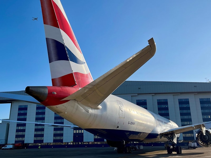 A British Airways airplane's tail in the foreground, with a hangar behind and another plane flying in the clear blue sky above.