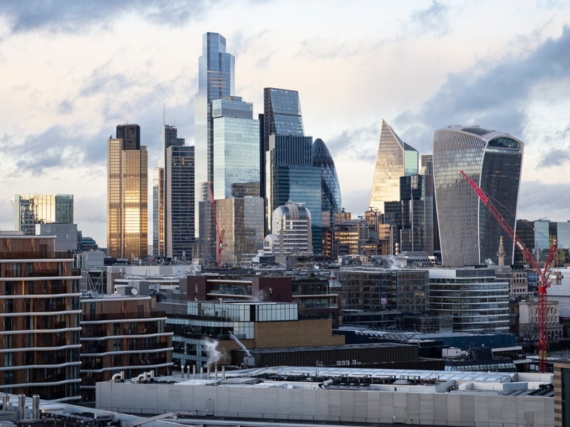 Panoramic view of the City of London skyline featuring modern glass skyscrapers, including the Gherkin, the Leadenhall Building and 20 Fenchurch Street (the Walkie Talkie). Residential and commercial buildings fill the foreground, while several red construction cranes are visible among the high-rise towers. 