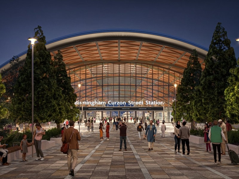 Profile view of the HS2 Curzon Street station’s exterior, showing the sweeping curved roof with timber-lined structure, large glass façade, and surrounding landscaped public spaces under a bright sky.