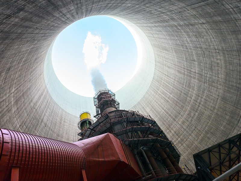 Upward view from inside a large industrial cooling tower, showing red and steel infrastructure rising toward a circular opening where steam vents into the sky above.