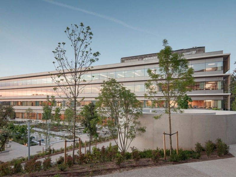 Modern three-story building surrounded by greenery, featuring large windows reflecting the sky at dusk. Pathway leads to the entrance.