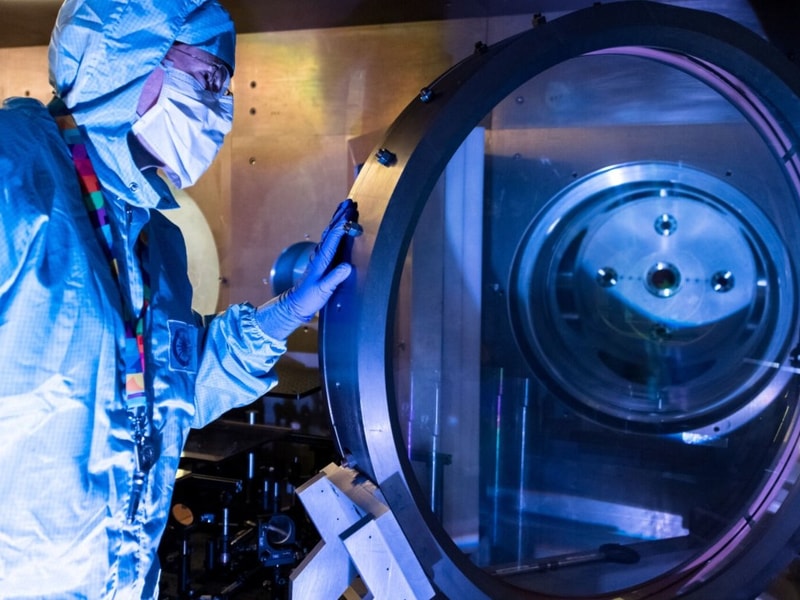 A person wearing full cleanroom protective clothing and face covering stands beside a large circular scientific instrument, looking through its transparent chamber. The scene is lit with blue and purple tones, and the equipment appears to be part of a high‑tech laboratory or manufacturing environment.
