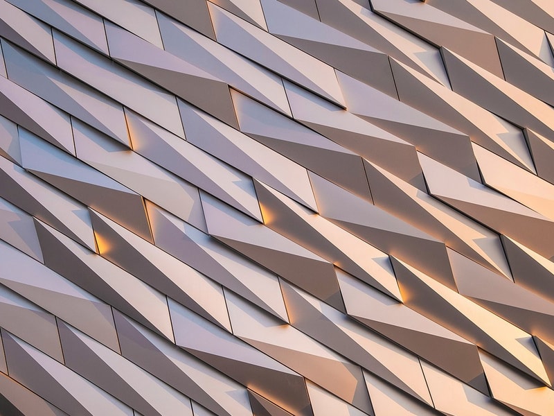 An abstract architectural façade made of angled metallic panels that catch soft light and create a repeating geometric pattern.