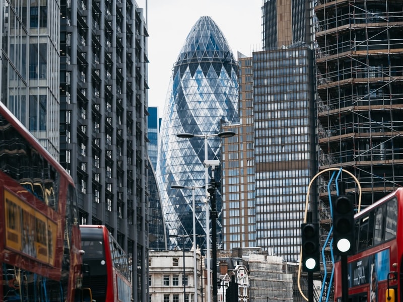 View along a busy London street lined with tall modern buildings, with the Gherkin skyscraper centred in the background. Red double-decker buses travel in both directions in the foreground, while a building on the right is covered in scaffolding. 