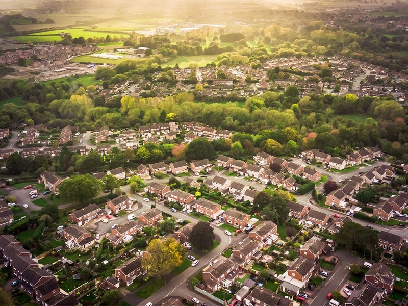 High-angle aerial shot of a suburban neighbourhood with rows of detached and semi‑detached houses surrounded by green trees and open countryside under soft evening light.