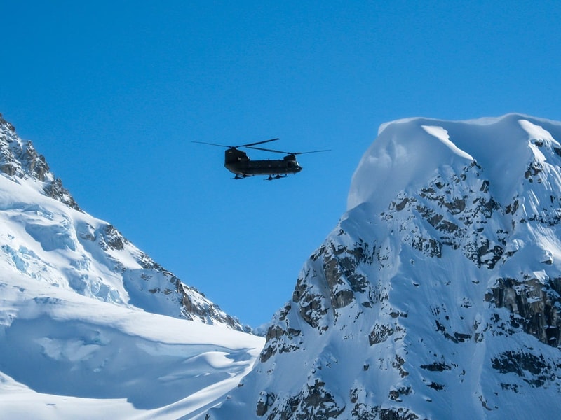 Helicopter flying between snow-covered mountain peaks under a clear blue sky.