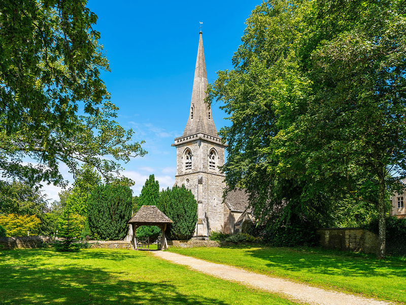 A stone church with a tall spire stands at the end of a grassy pathway, surrounded by large trees and bright greenery under a clear blue sky.