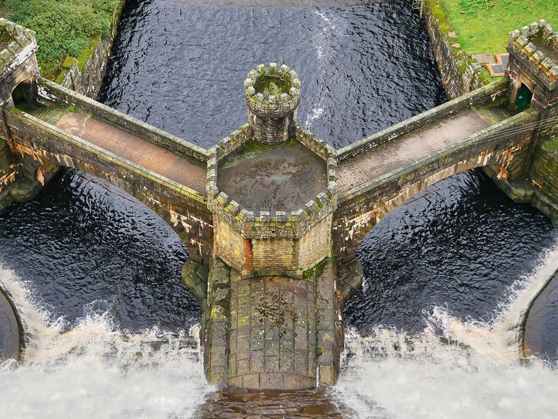Aerial view of a historic stone dam spillway with symmetrical walkways and turrets, directing water through multiple channels surrounded by green landscape.