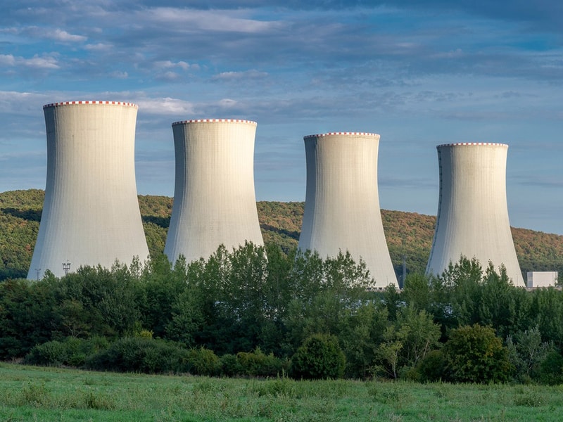 Four large cooling towers of a nuclear power plant rise behind a green forested area with hills in the background under a partly cloudy sky.