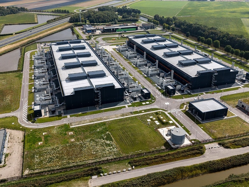 An aerial view of a large industrial facility with multiple long rectangular buildings, service roads, and surrounding fields in a rural landscape.