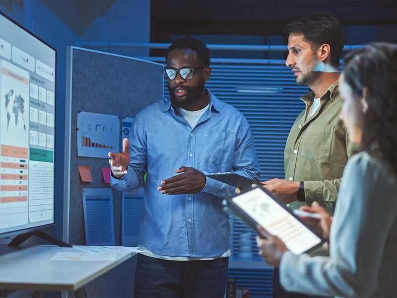 A group of people in an office setting review data visualizations on a large screen while discussing analytics and using a digital tablet.