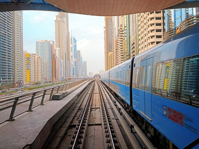 Blue metro train travelling along elevated tracks through a dense modern cityscape of high-rise buildings, viewed from a central track perspective under a station canopy.