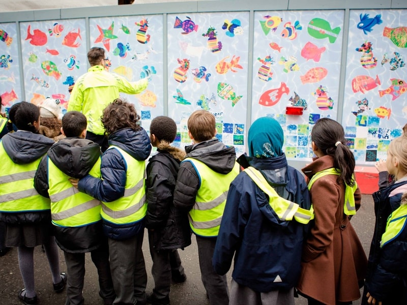 A group of children in high‑visibility vests stand in front of a colourful wall mural featuring fish artwork as an adult points out details.