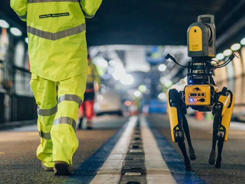 A worker in high-visibility yellow protective clothing walks alongside a four-legged robotic scanning device on a railway platform or track inspection area. The robot, equipped with sensors and cameras, moves along the platform edge under bright station lighting, with rail infrastructure blurred in the background.