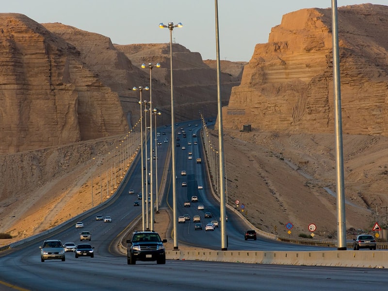 A multi-lane highway carrying steady traffic winds through a dramatic desert landscape, with tall streetlights lining the road as it passes between towering sandstone cliffs under warm evening light.