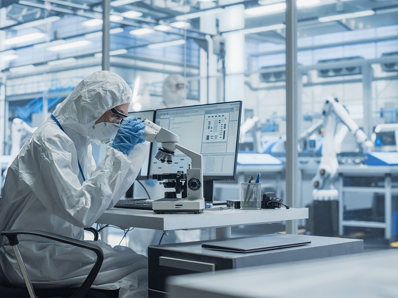 A person in full protective laboratory gear working with a microscope at a workstation equipped with a computer inside a high-tech industrial lab environment.