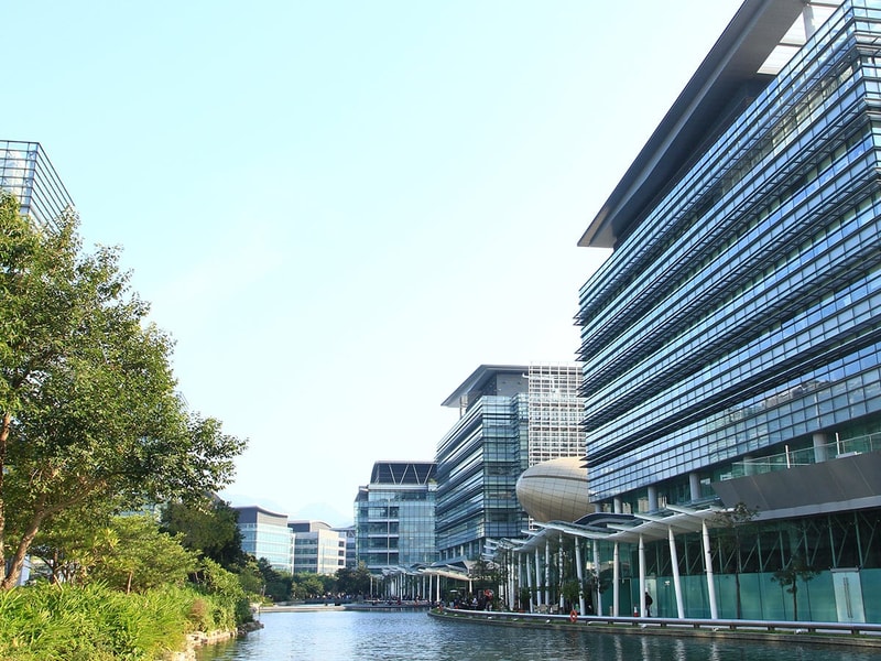 A collection of contemporary glass office buildings lining a landscaped waterfront, with trees, reflective waterways and modern architecture creating a business district setting.