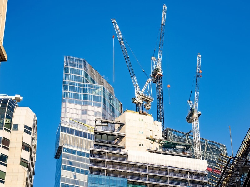 A construction site with cranes and partially built high‑rise structures stands against a clear blue sky in a modern urban area.