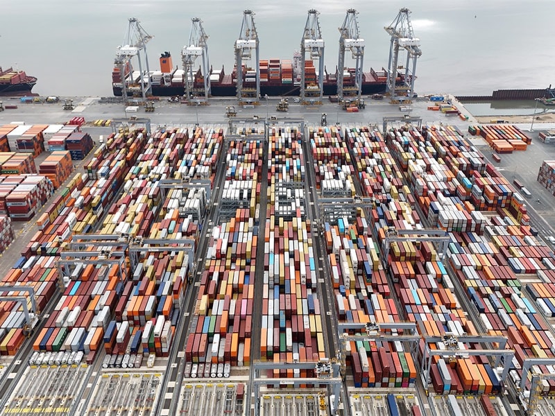An aerial view of a large container port showing rows of multicoloured shipping containers stacked in long grids, with cranes lining the quay and cargo ships docked beside the water.