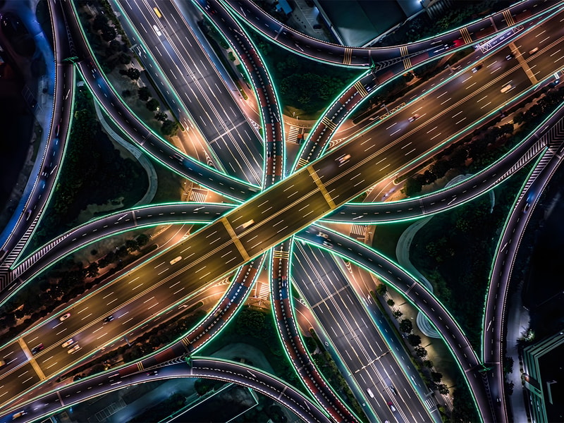 An aerial view of a brightly illuminated motorway interchange at night shows intersecting highways, flowing traffic, and illuminated roadways forming a complex transport network.