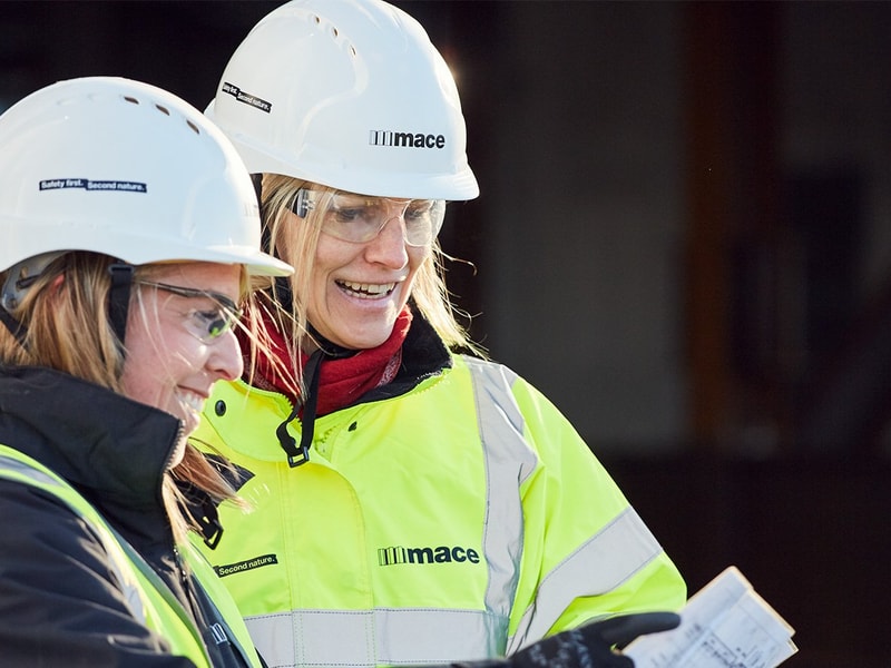 Two construction workers in hard hats and high-visibility jackets are reviewing plans on a clipboard at a construction site.