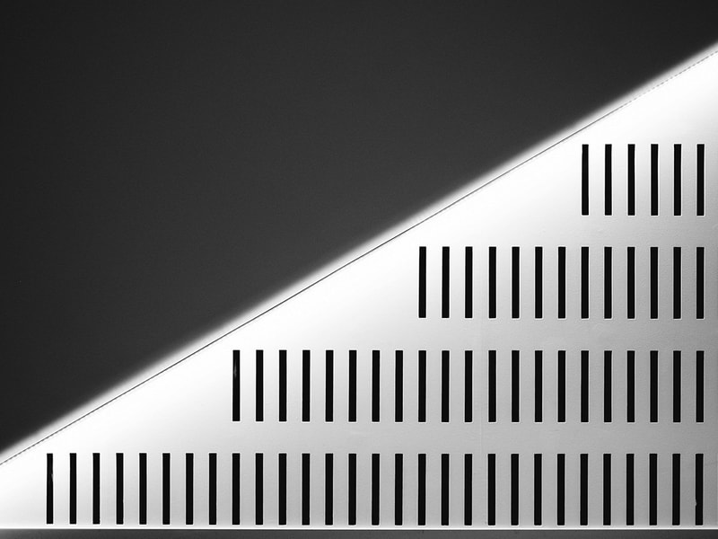 A minimalist black‑and‑white architectural detail shows diagonal lighting cutting across a wall of evenly spaced vertical slits.